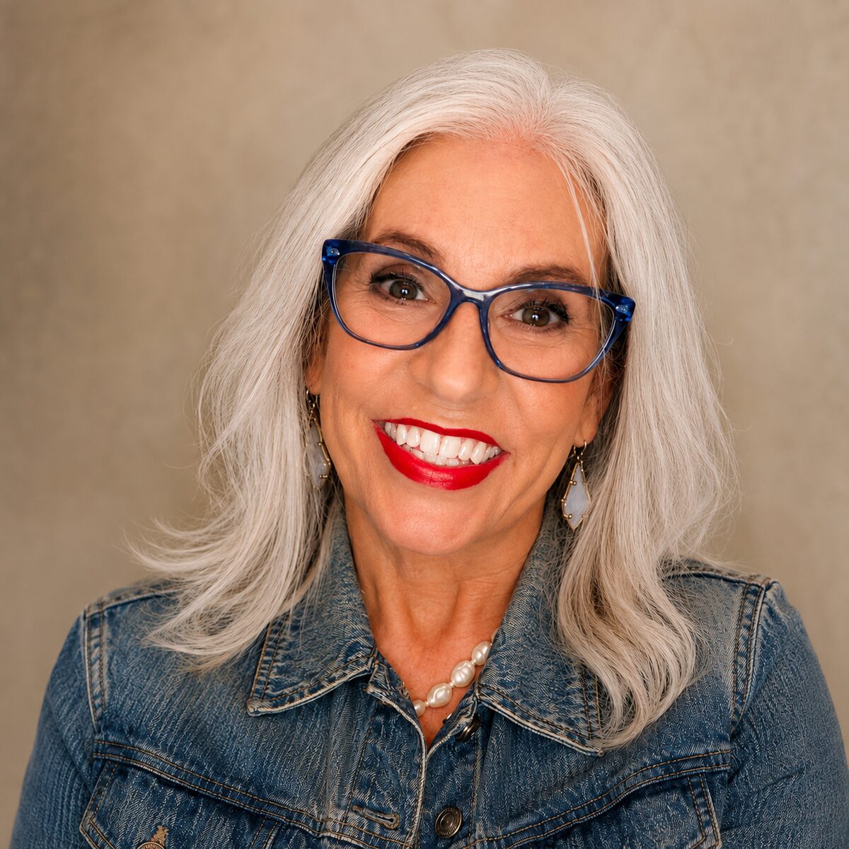 Portrait of Robbyn Andersen, smiling, with long silver hair, blue glasses, and a denim jacket